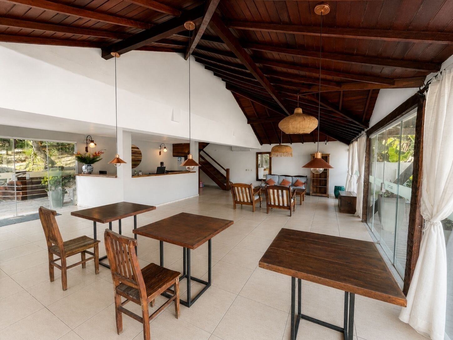 Um espaço amplo e interno, com mesas e cadeiras de madeira, um balcão de recepção e uma escada que leva a um andar superior.