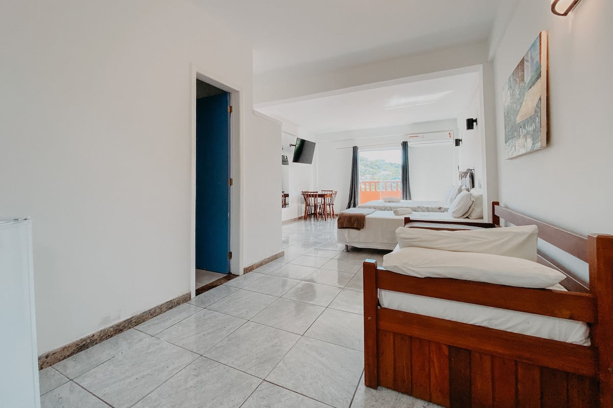 Quarto de hotel moderno com cama de madeira, sofá, área de refeições com mesa e cadeiras, e uma grande janela com vista.