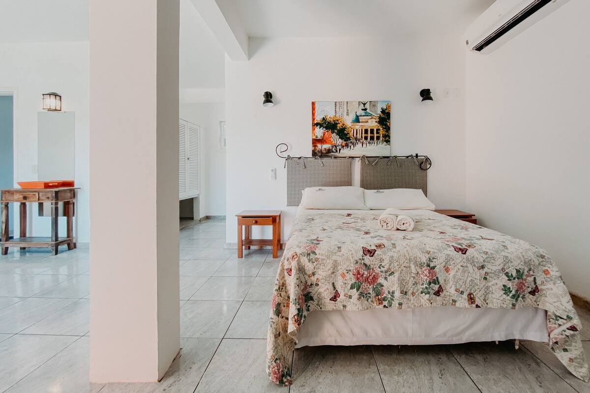 Quarto com cama grande, uma mesinha, uma cadeira e um quadro na parede.