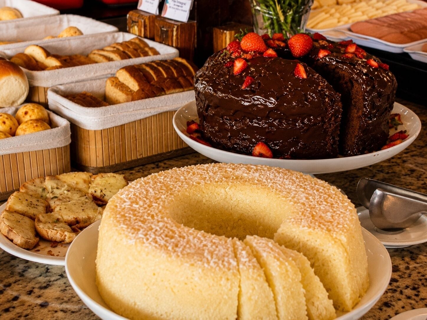 Un bufé con diversas opciones de panadería, incluyendo pan, bollos y un pastel de chocolate con fresas.