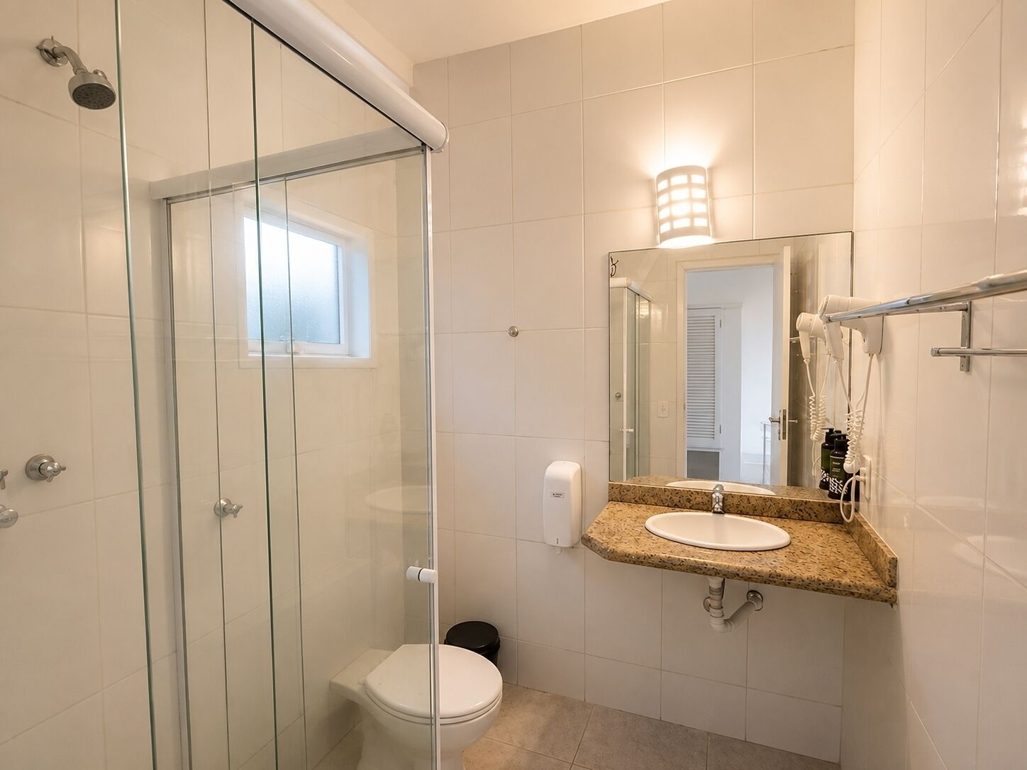 Baño con ducha envuelta en vidrio, inodoro, lavabo con espejo y secador de pelo en la pared.