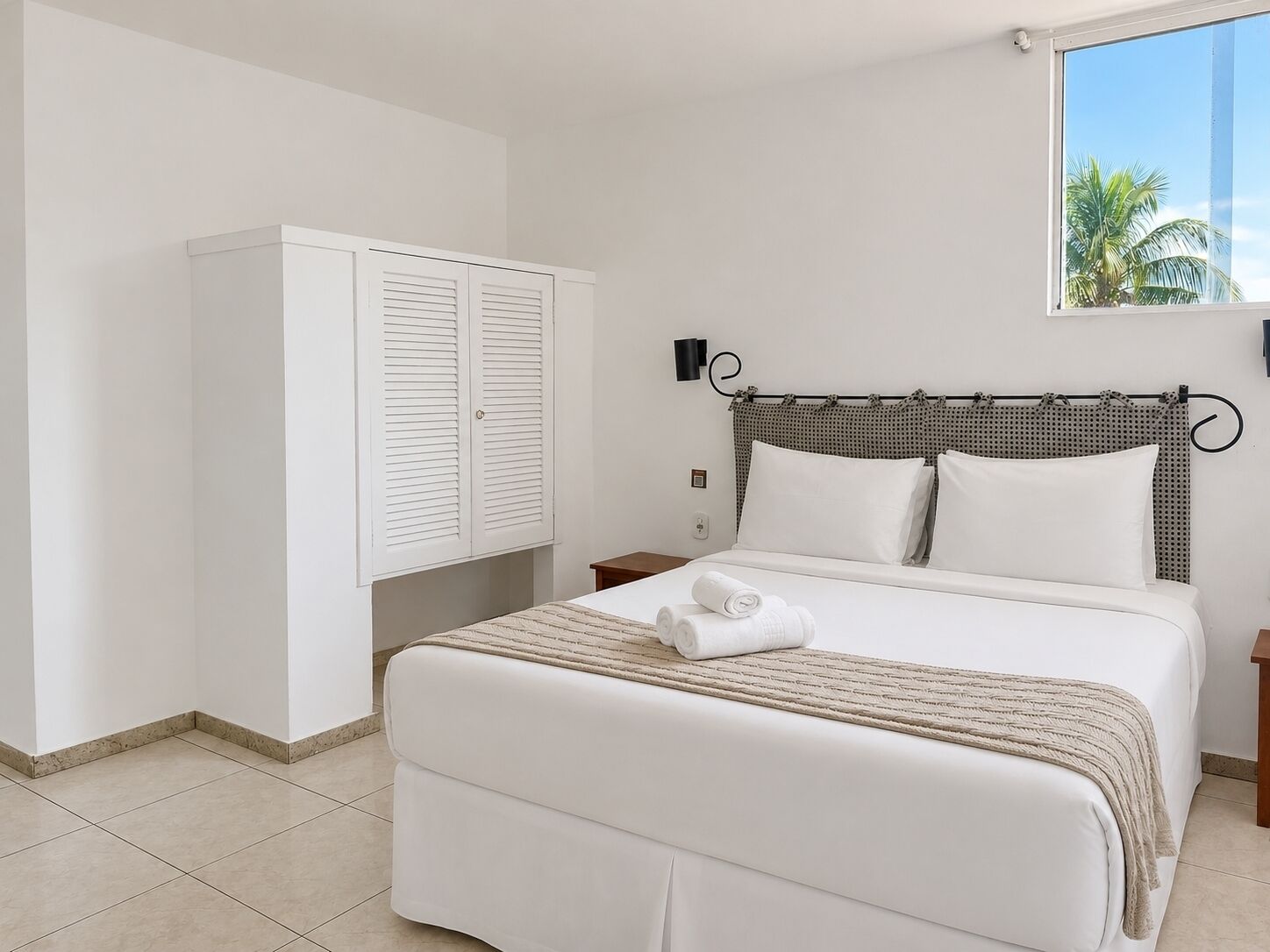 Habitació d'hotel amb un llit, un armari, una tauleta de nit i una finestra amb vista a palmeres.