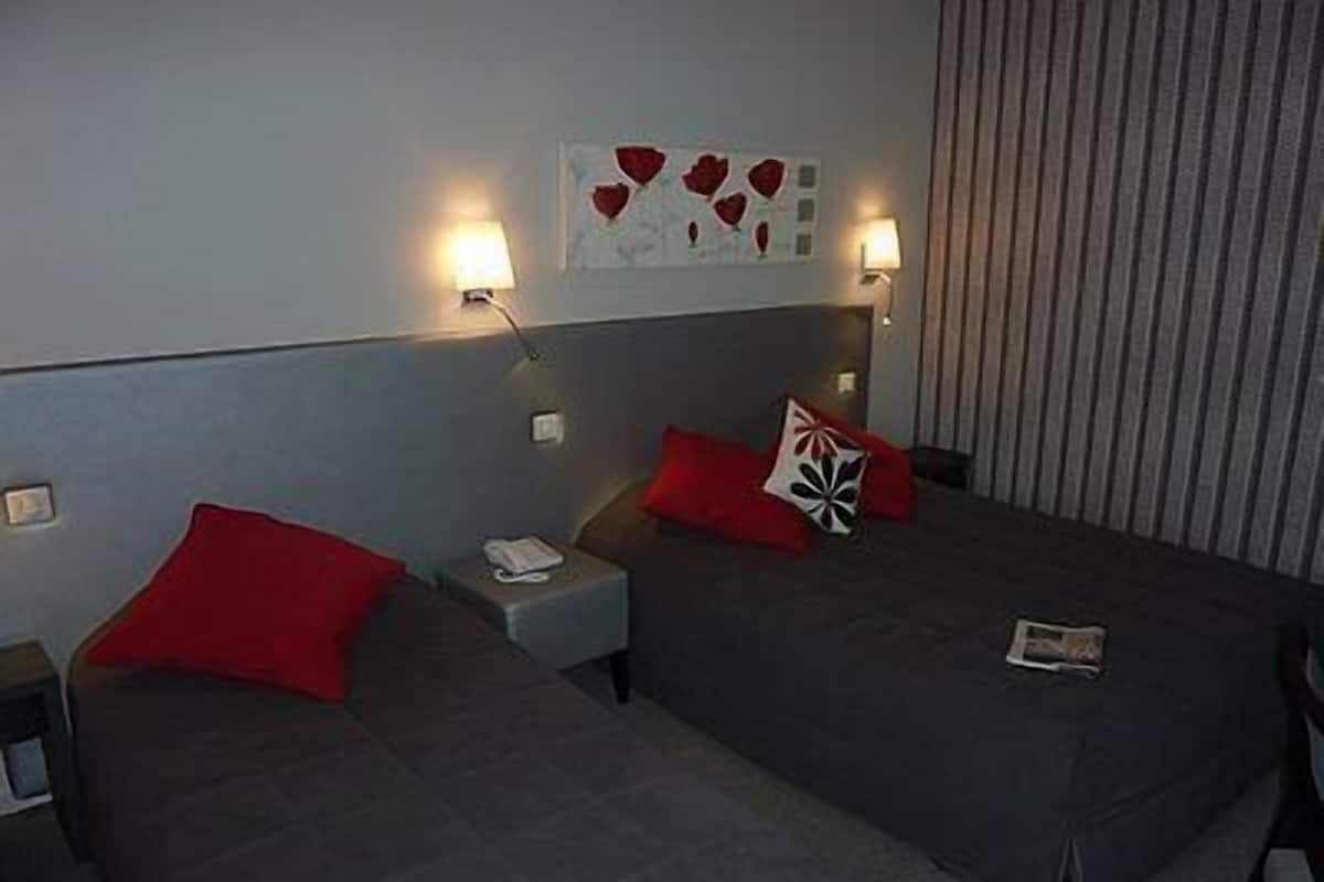 A hotel room with two beds, red pillows, a nightstand with a phone, and a wall art piece.