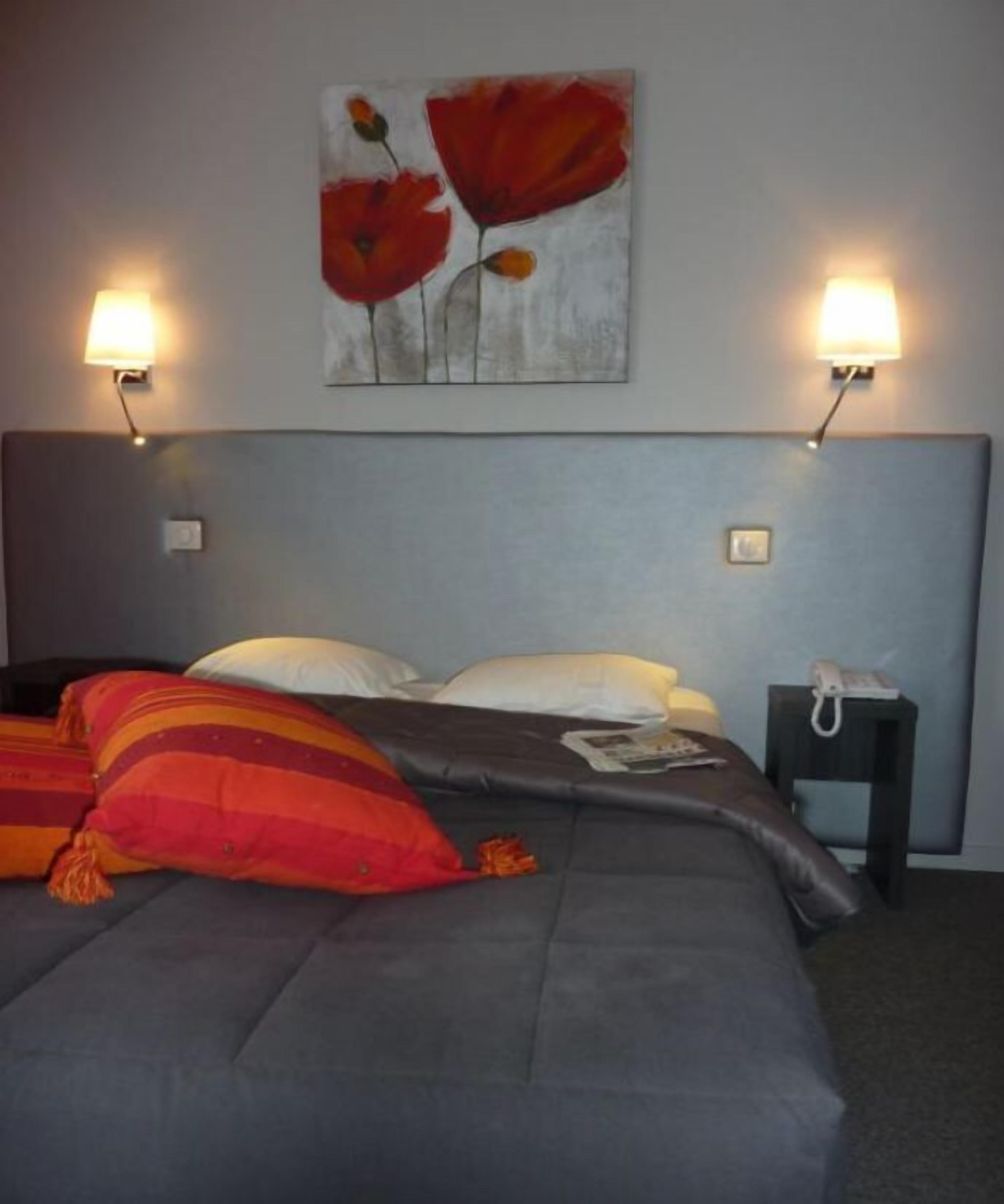 A bed with grey bedding, red and orange pillows, and a painting of red flowers on the wall.