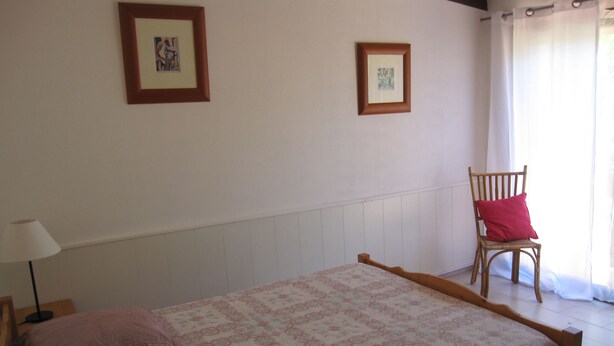 Quarto com cama, duas almofadas, uma cadeira e duas imagens emolduradas na parede.