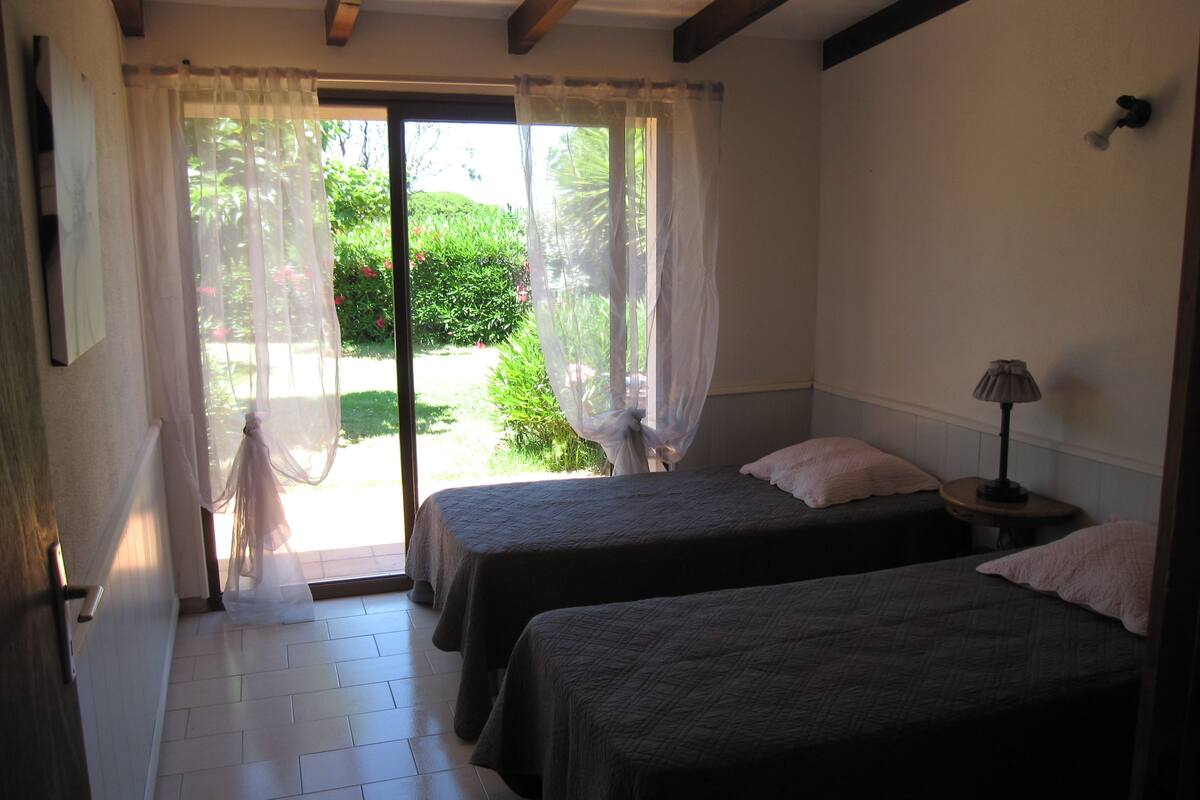 A room with two beds, a small table with a lamp, and a view of greenery outside.