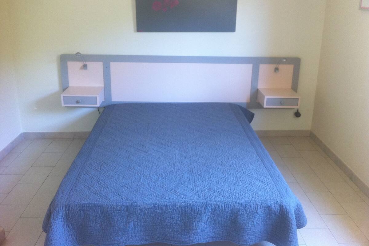 A bed with a blue bedspread, a headboard with two bedside tables, and a wall-mounted shelf above.