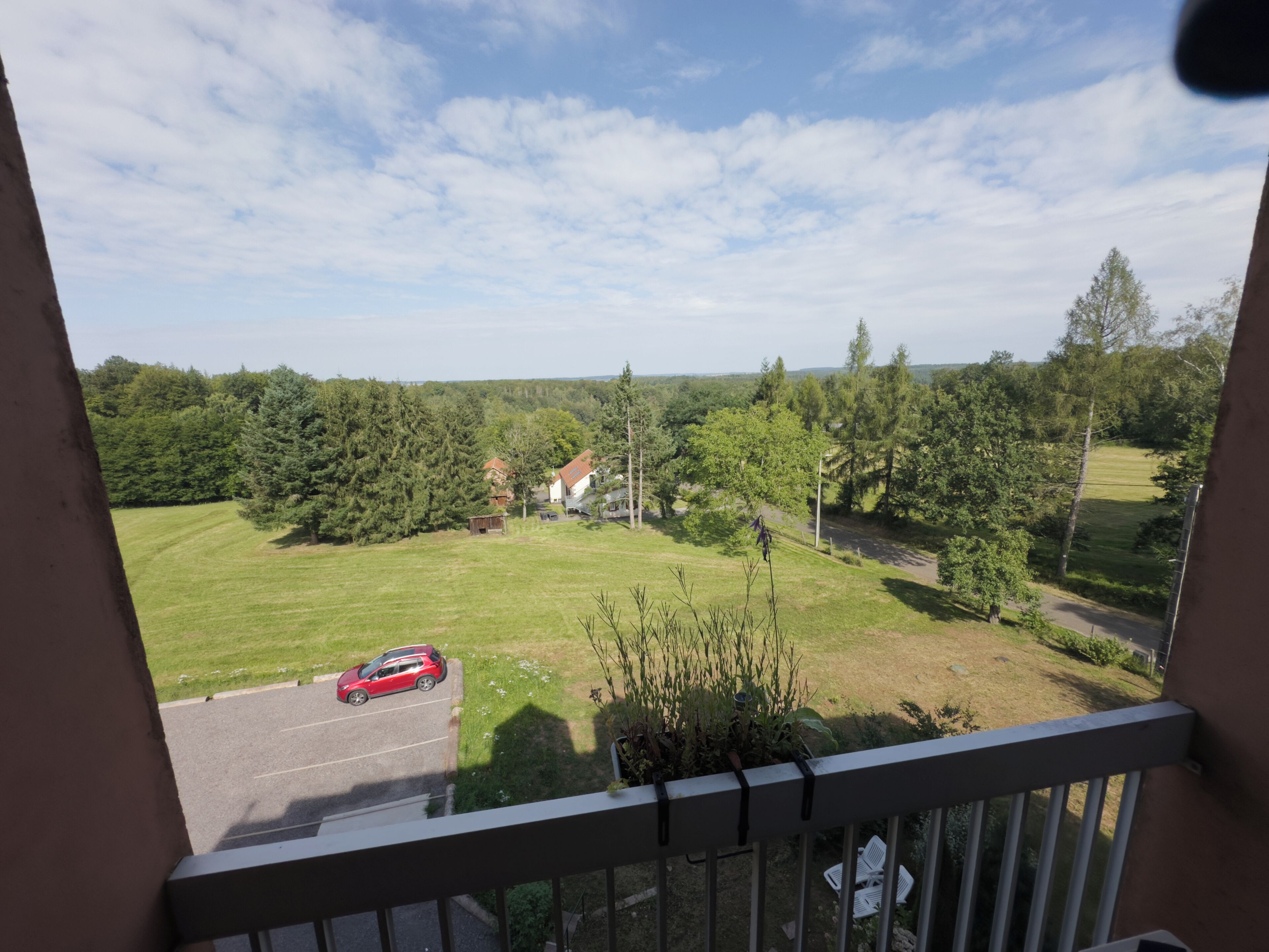 Ein Balkon mit Blick auf ein grünes Feld, Bäume und ein rotes Auto auf der Straße.