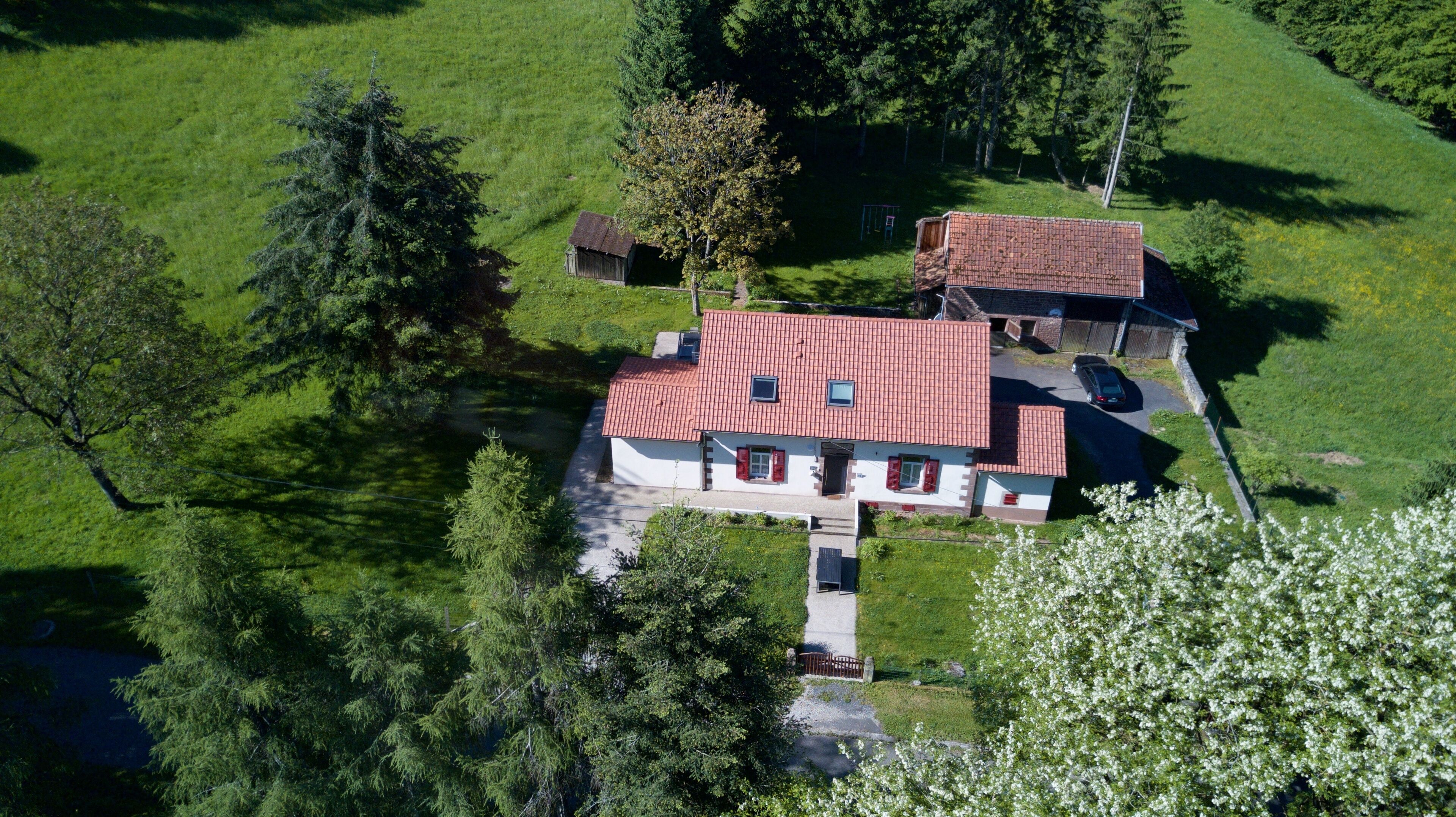 Une maison à deux étages, avec un toit recouvert de tuiles rouges, entourée de champs verdoyants et d’arbres.
