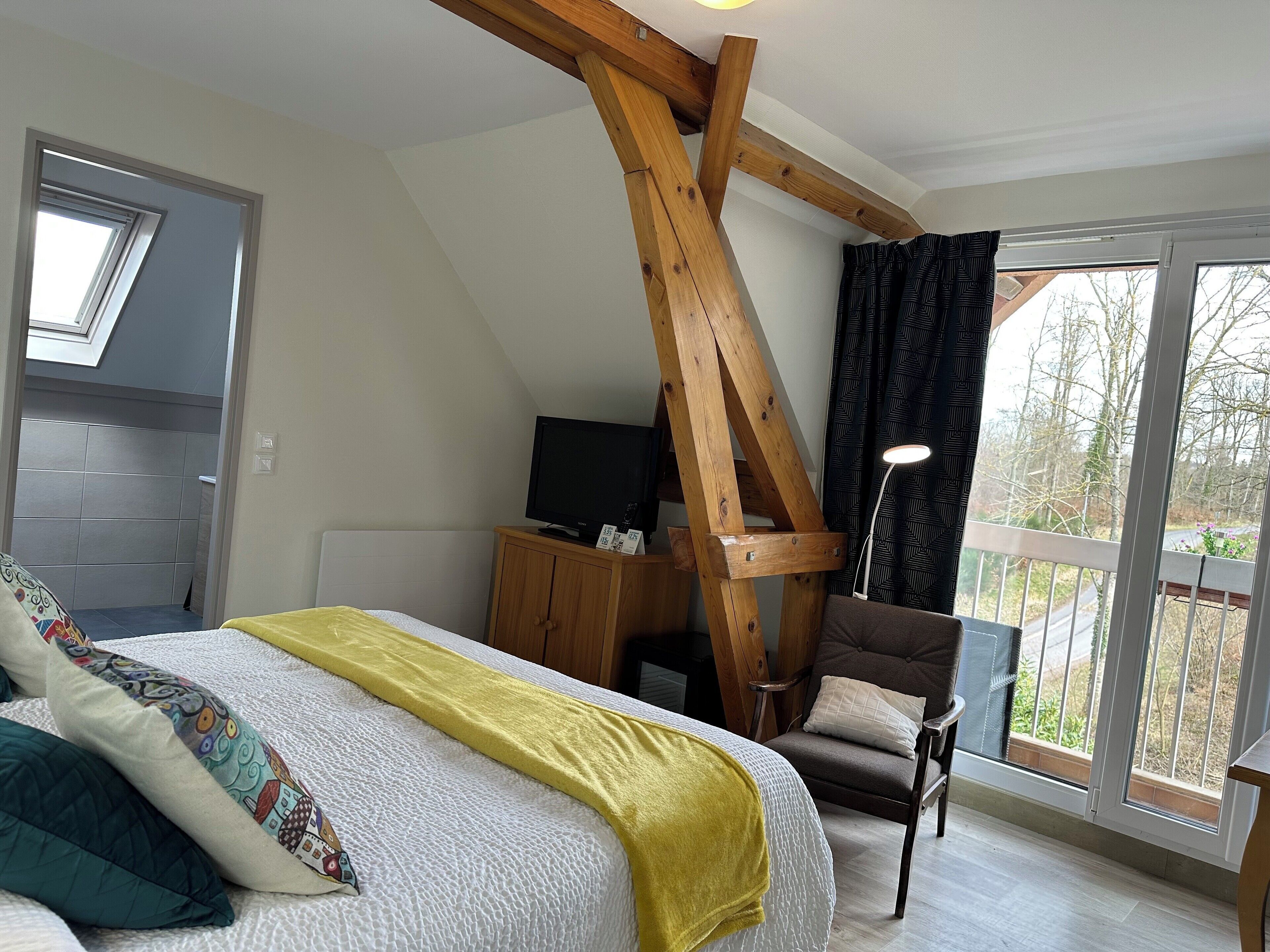 Un dormitorio con techo inclinado, una cama con una manta amarilla, una silla de madera, un televisor sobre un mueble de madera y vistas a árboles a través de una ventana.