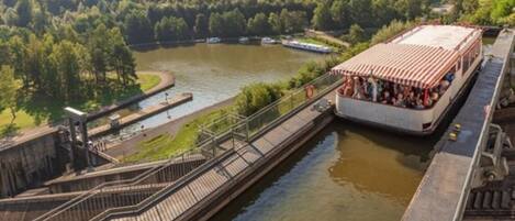 A riverboat docked at a lock system with a bridge and forested hills in the background.