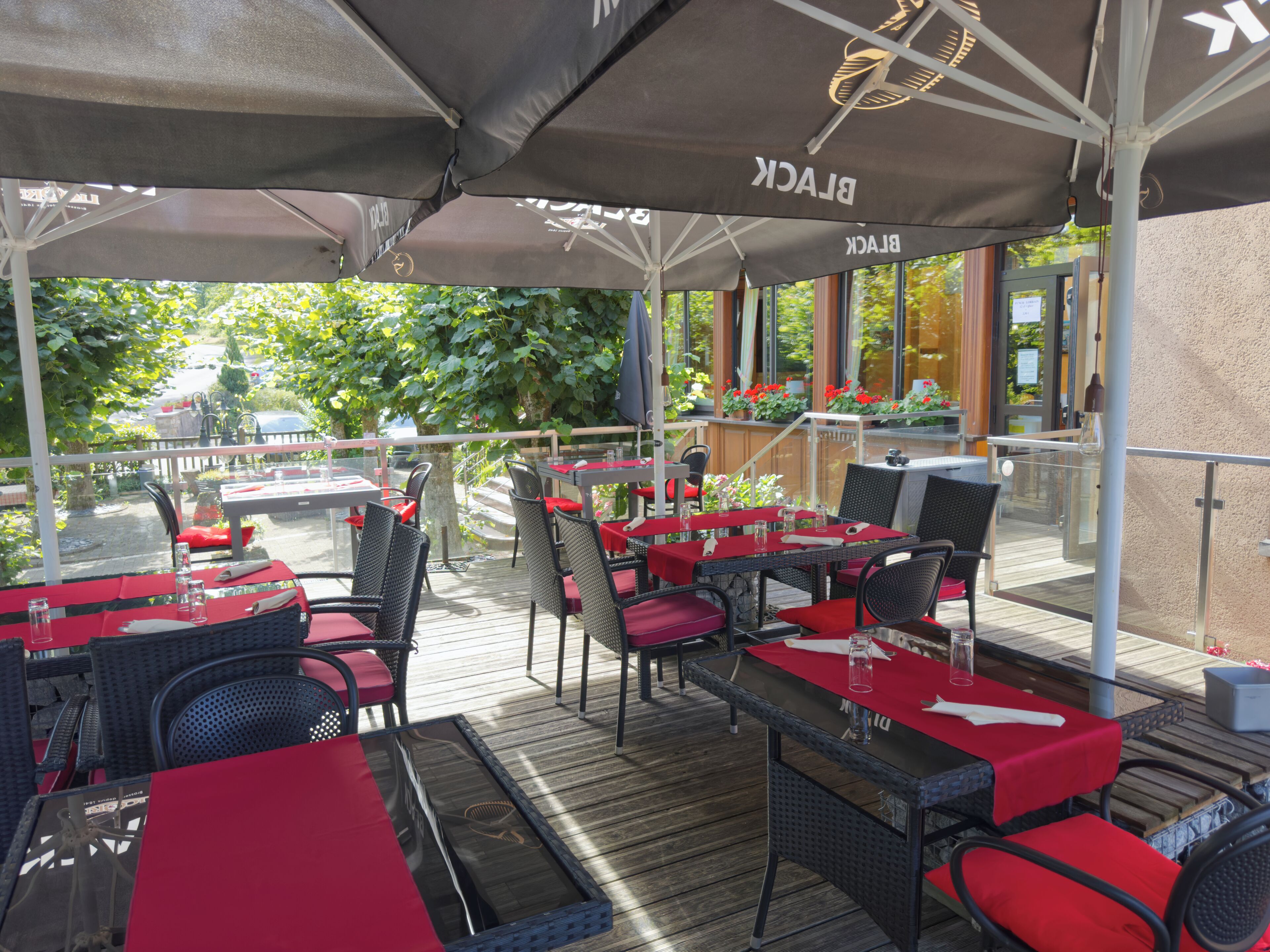 An outdoor patio with tables and chairs, covered by umbrellas, and a view of the street and greenery.