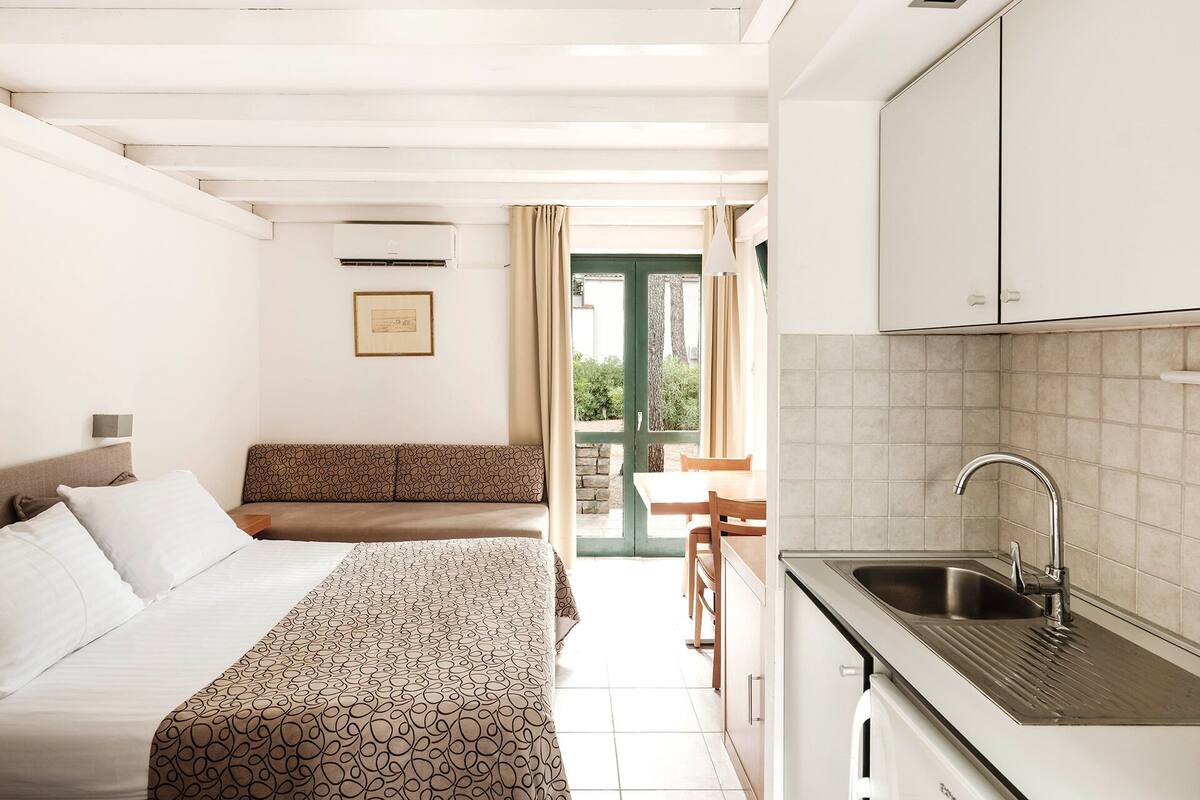 A hotel room with a bed, a sofa, a dining table, and a kitchen area with a sink.