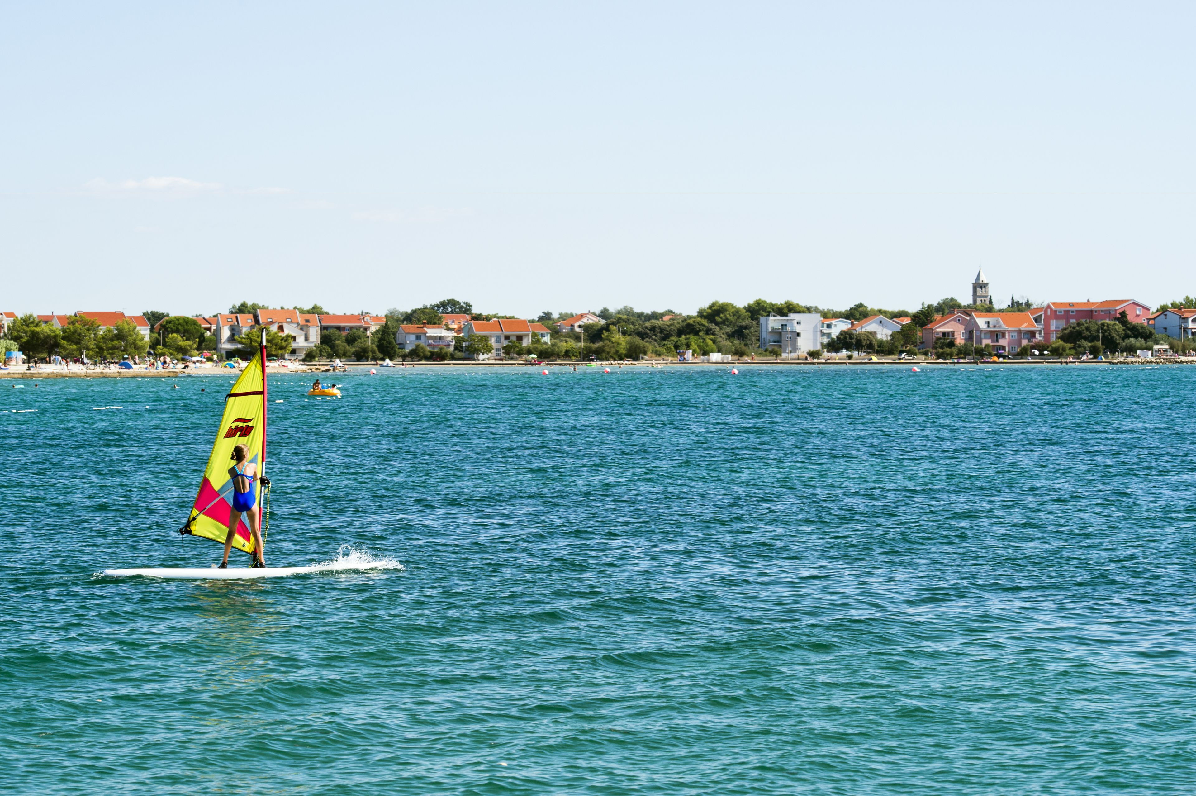 Une personne pratiquant la planche à voile sur une étendue d’eau, avec des bâtiments en arrière-plan.