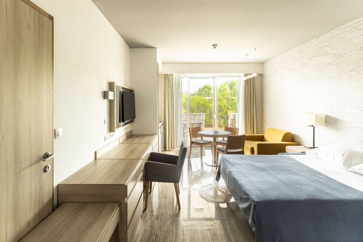 A hotel room with a bed, a desk, a chair, a small table, and a TV.