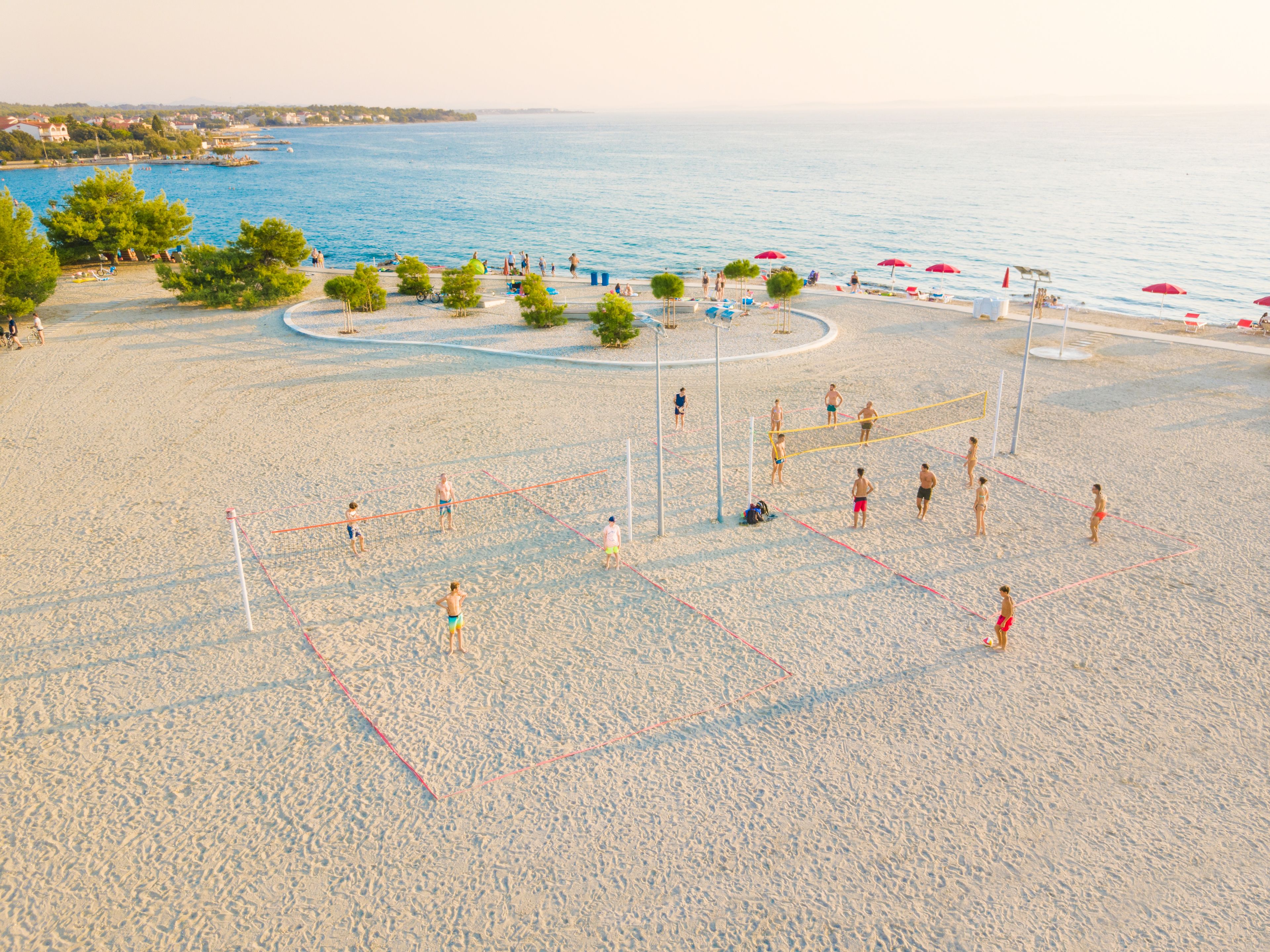 A beach volleyball court with players and spectators, surrounded by trees and beach umbrellas.