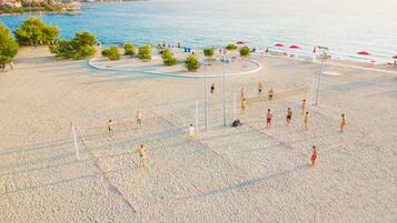 Cancha de voleibol de playa con jugadores y espectadores, rodeada de árboles y sombrillas.