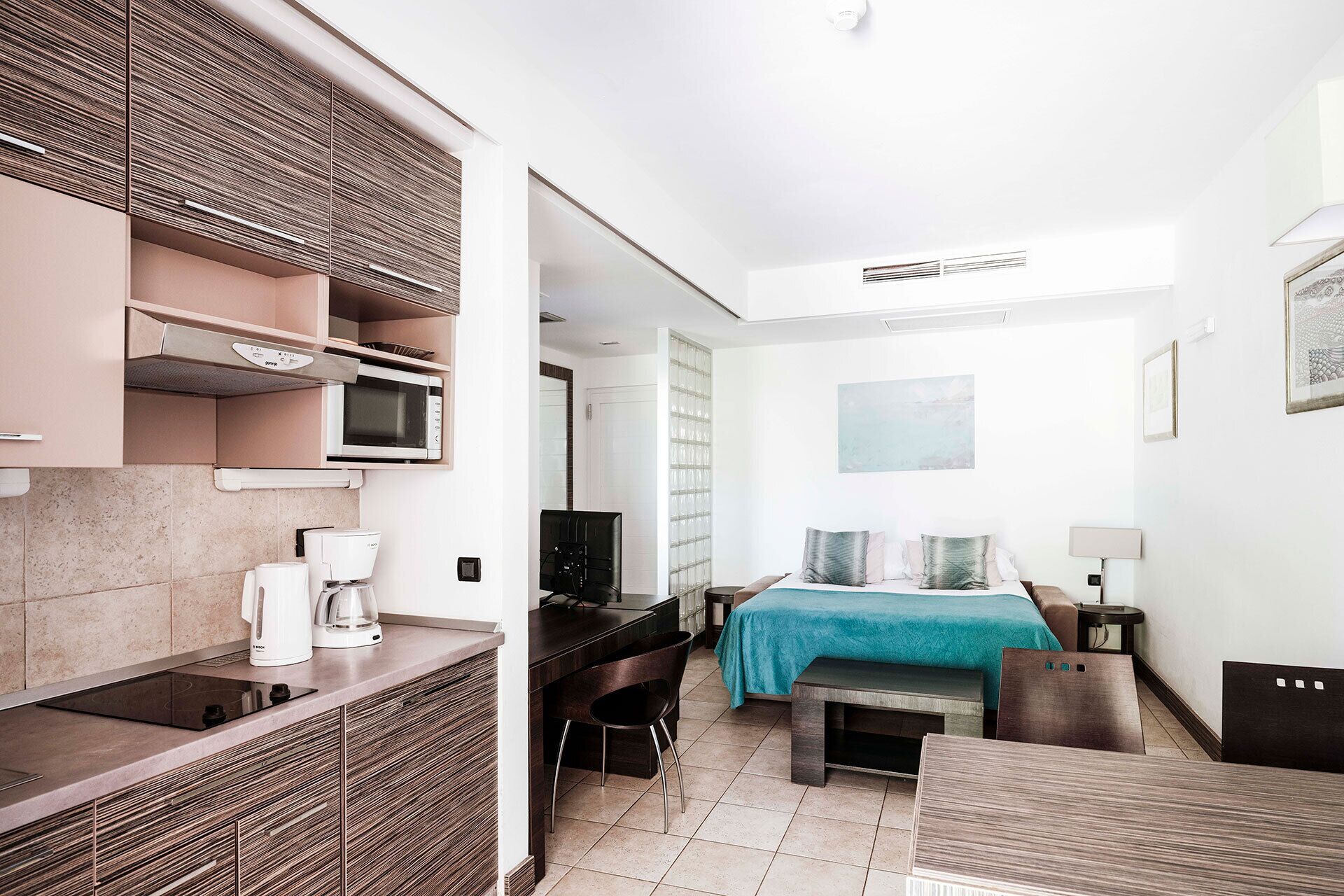 Een compacte hotelkamer met kitchenette, een bed met een blauwgroene sprei, een bureau, een stoel en een nachtkastje.