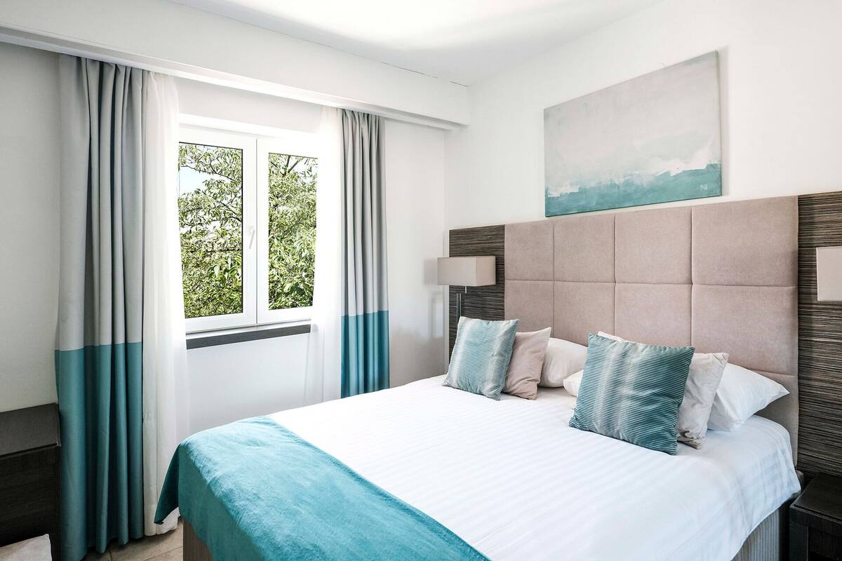 A hotel room with a bed, curtains, a window with a view of trees, a headboard, and a painting on the wall.