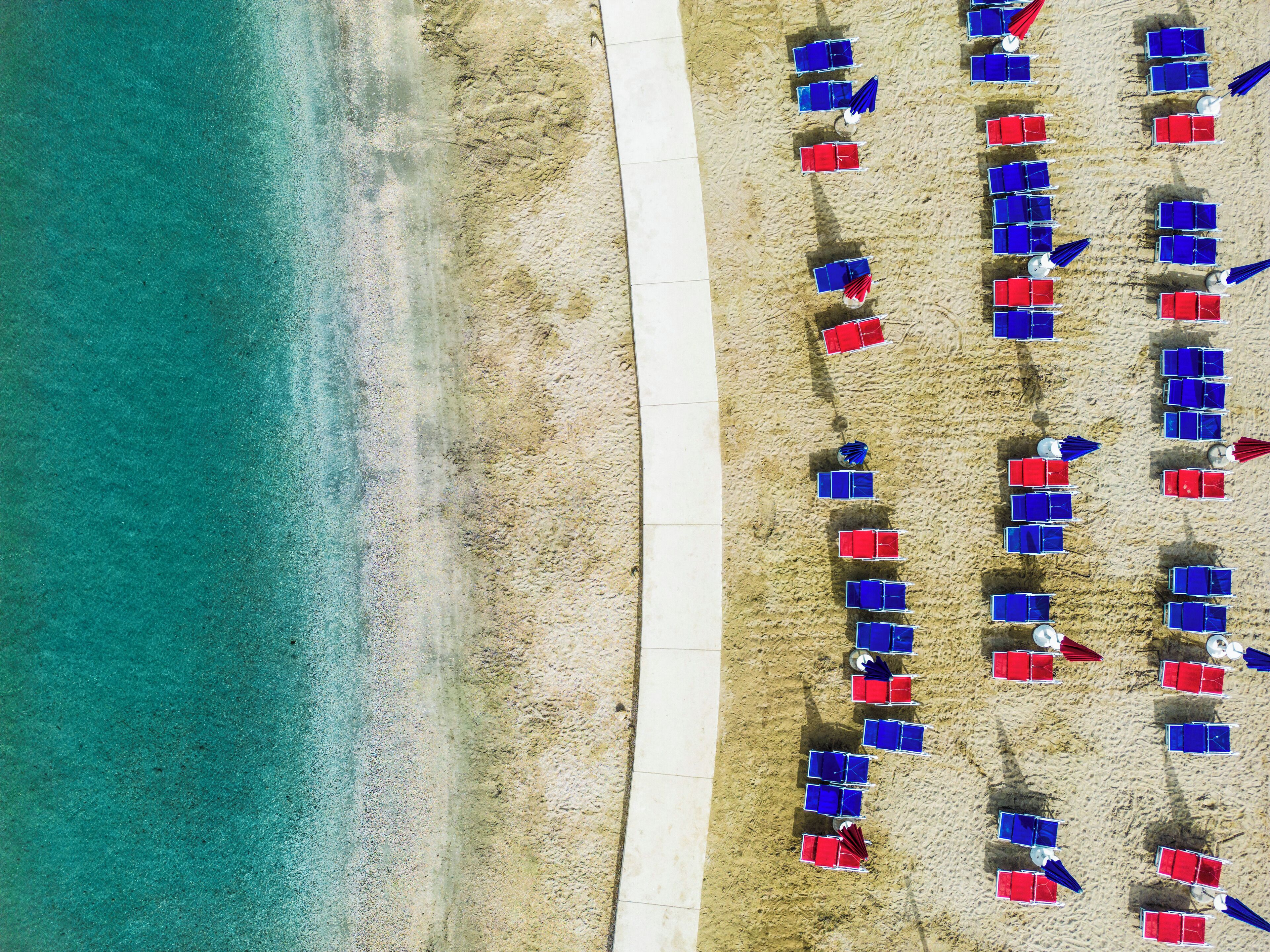 Vista aérea de una playa con filas de sillas y sombrillas azules y rojas alineadas a lo largo de la costa.