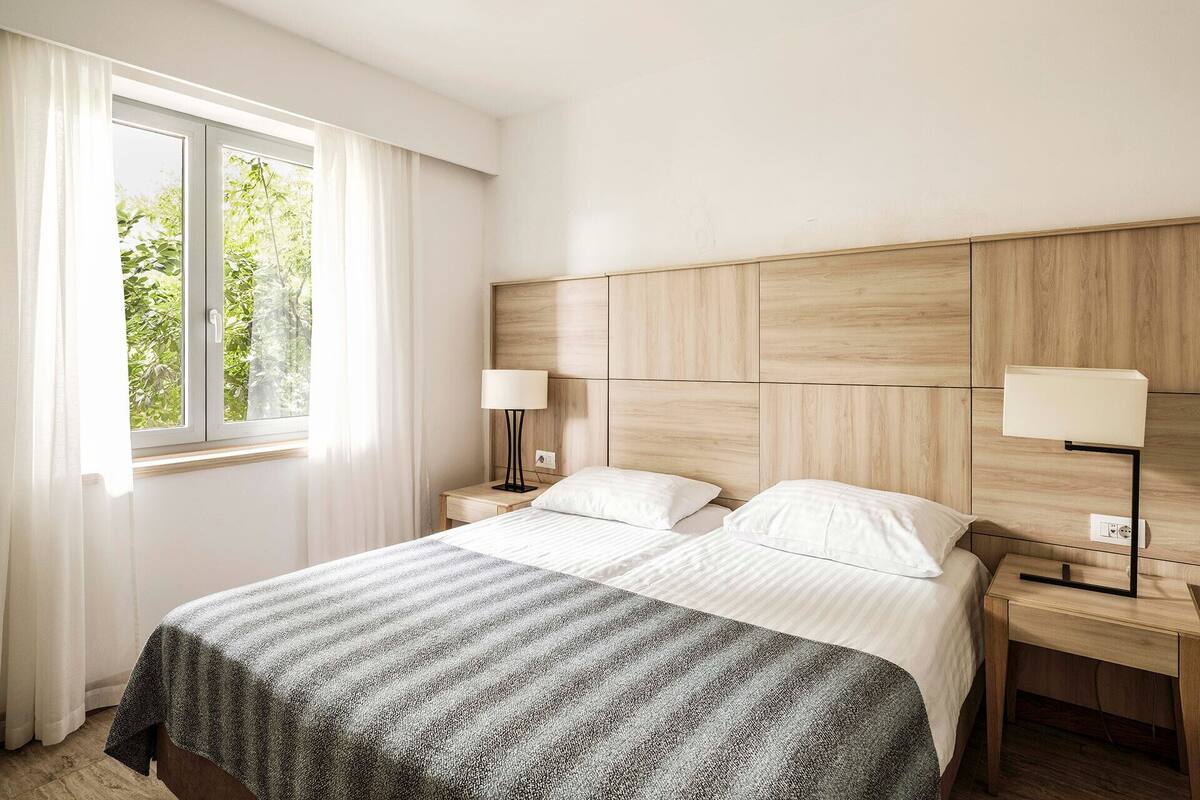 A hotel room with a large bed, two bedside tables, a window with curtains, and a view of greenery outside.