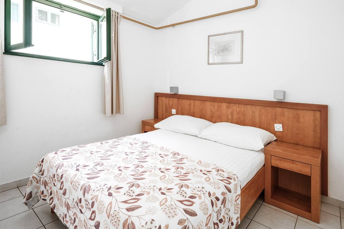 A bedroom with a wooden headboard, a bed with white linens, a nightstand, a bedside table, a window with curtains, and a framed picture on the wall.