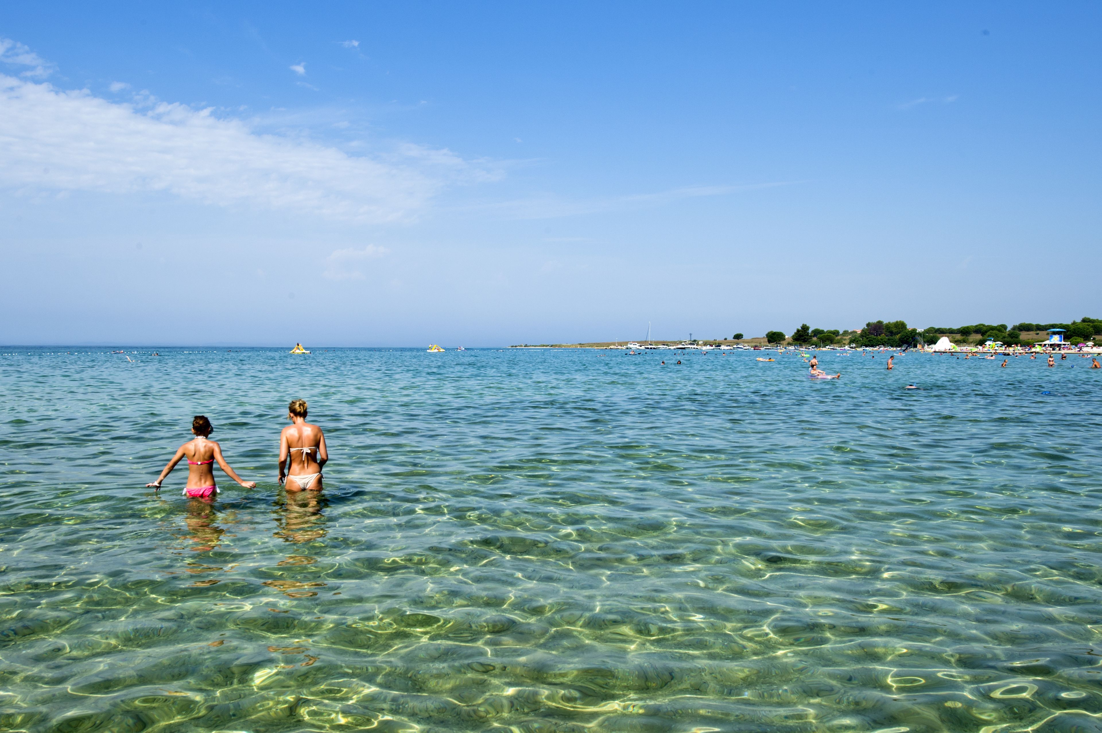 A beach with clear water, people swimming, and a distant shoreline with buildings.