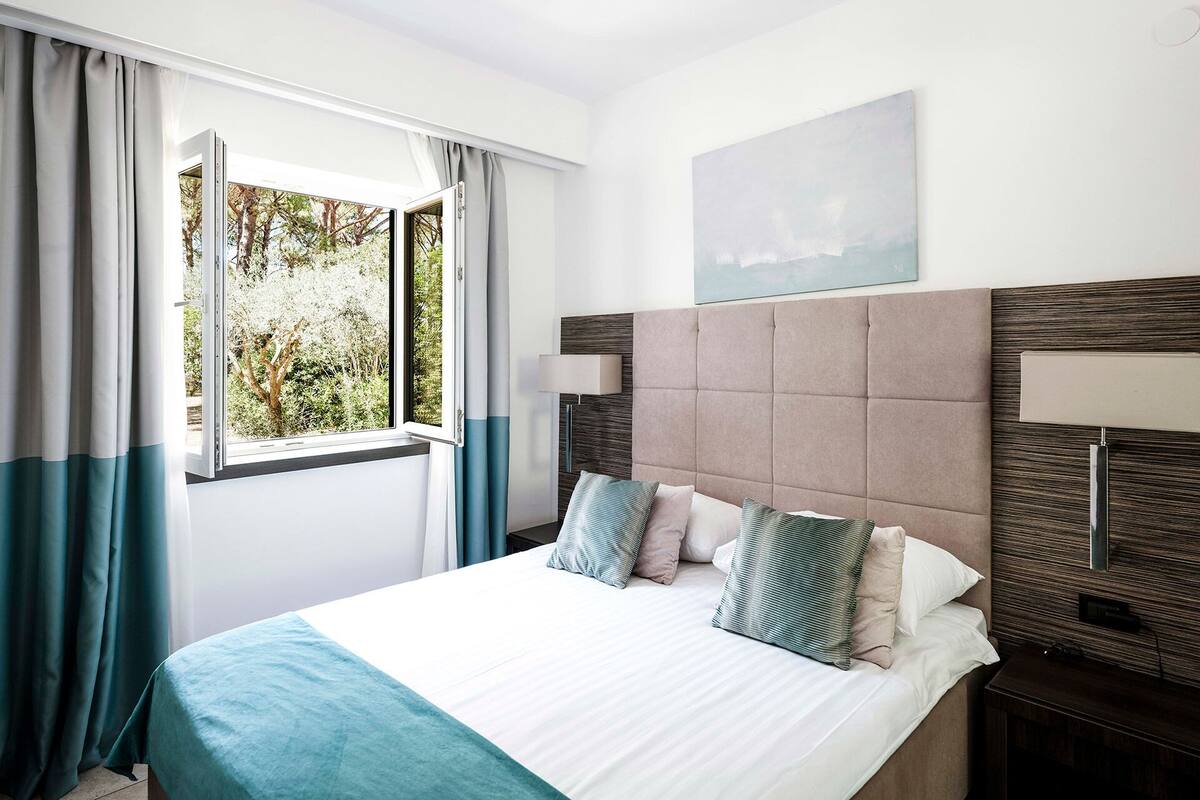 A hotel room with a bed, a window with curtains, a headboard, a bedside table, and a wall-mounted lamp.