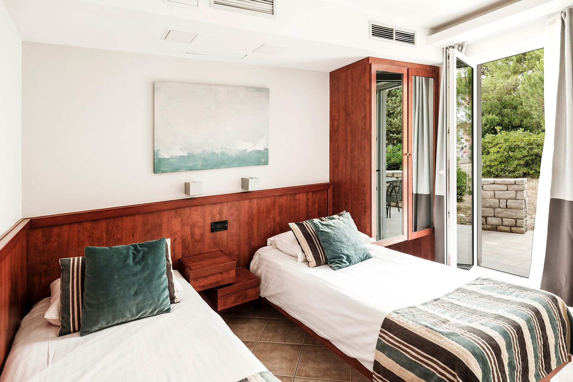 Habitación de hotel con dos camas, cabecero de madera, un cuadro en la pared y vistas a la vegetación a través de una puerta corredera.