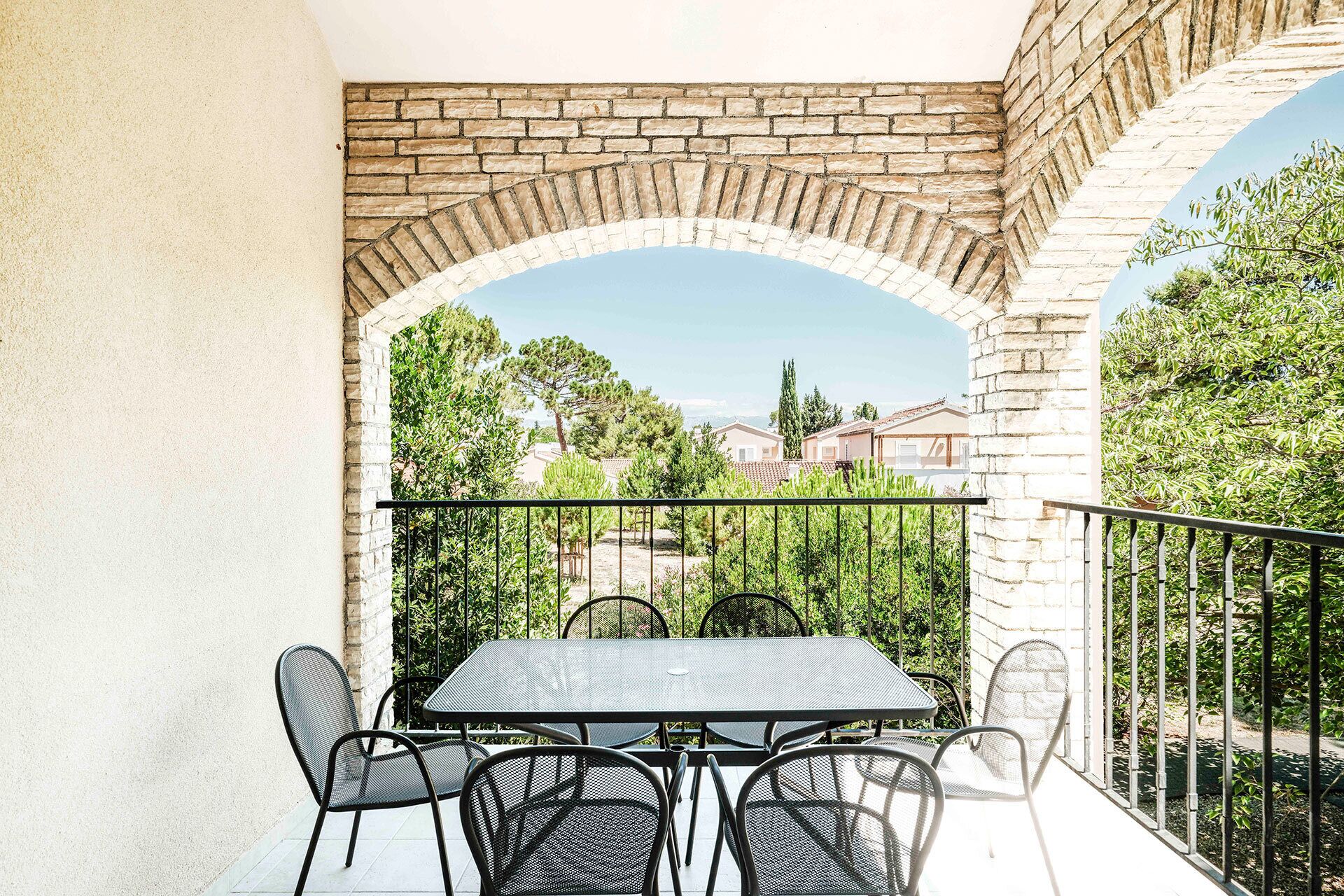 A balcony with a table and chairs, a stone archway, and a view of houses and trees.