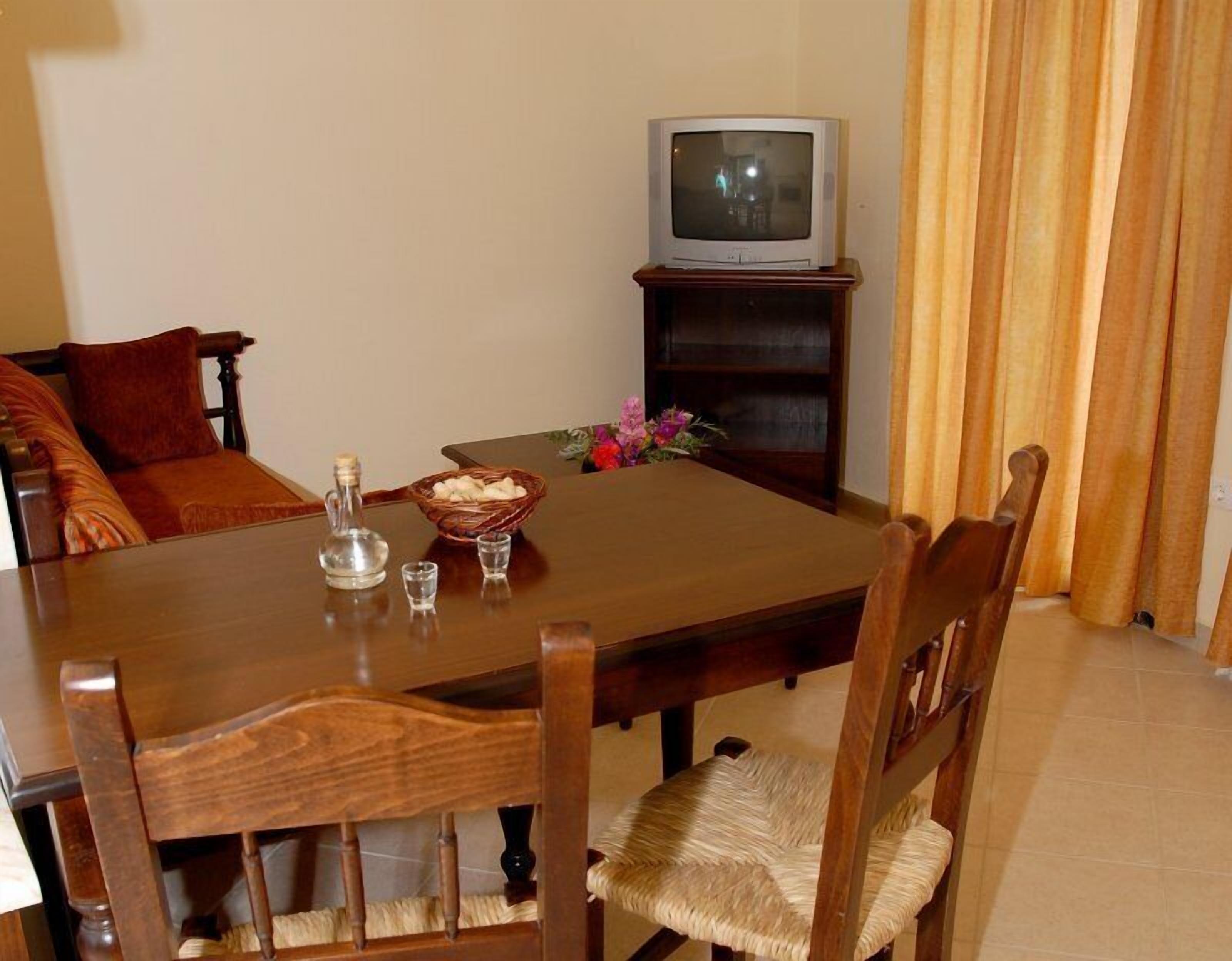 A dining area with a wooden table, chairs, a television on a stand, a vase with flowers, and a bottle.