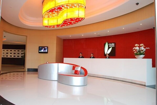 A hotel reception area with a modern design, featuring a curved reception desk, a large chandelier, and a red accent wall with the hotel's logo.