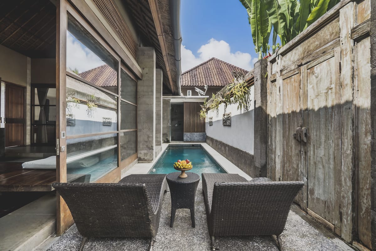 A pool area with wicker chairs and a table, a wooden fence, and a building with a tiled roof.