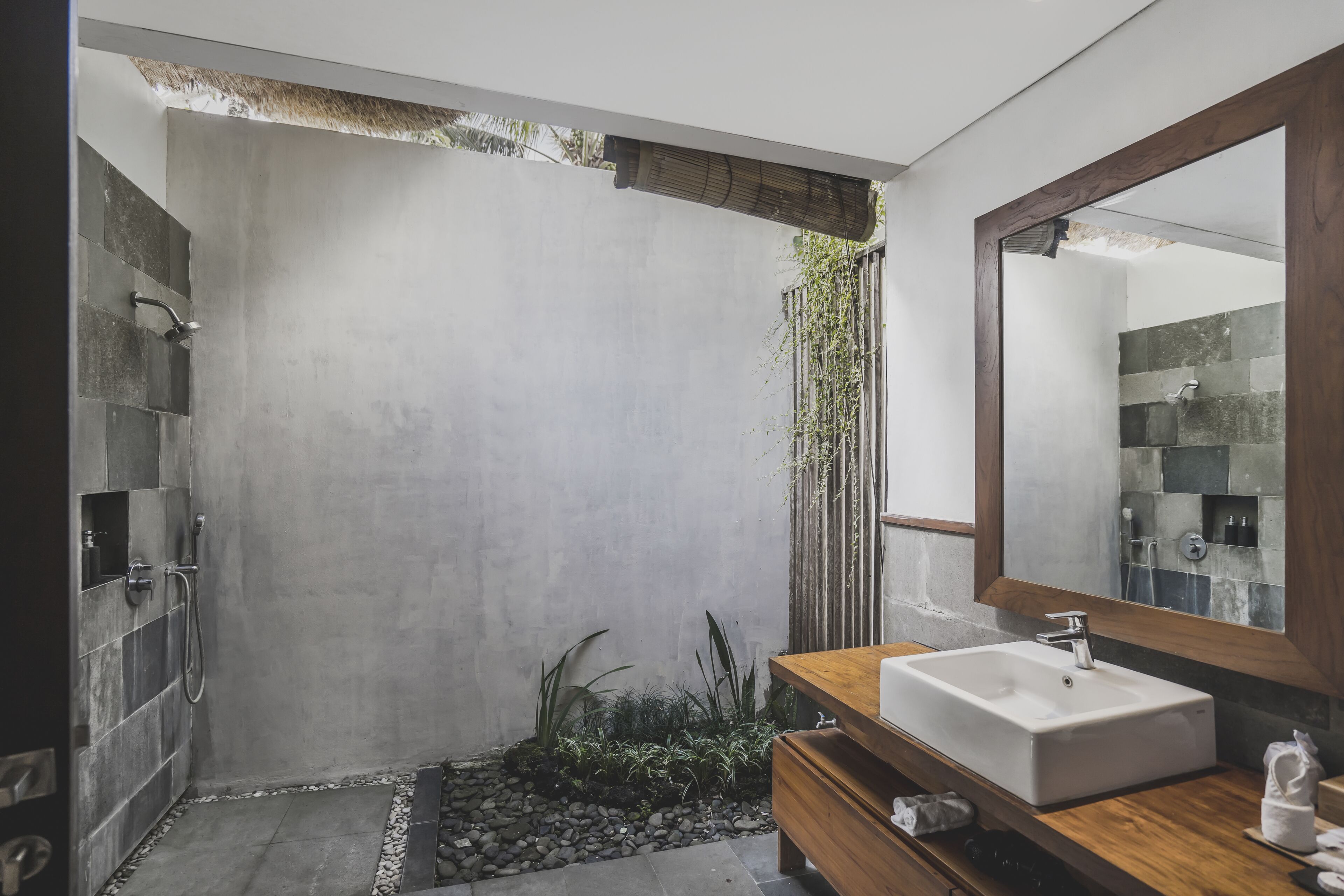A modern bathroom with a large mirror, a wooden vanity, and a shower area with a concrete wall and plants.