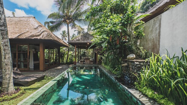 A swimming pool with a stone-lined edge, surrounded by lush greenery and a thatched-roof structure. - Luwak Ubud Villa & Jungle Suite (Ubud)