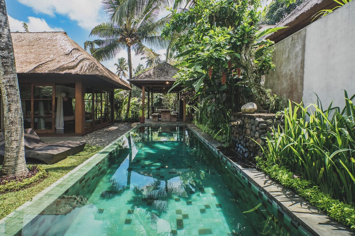 A swimming pool with a stone-lined edge, surrounded by lush greenery and a thatched-roof structure.