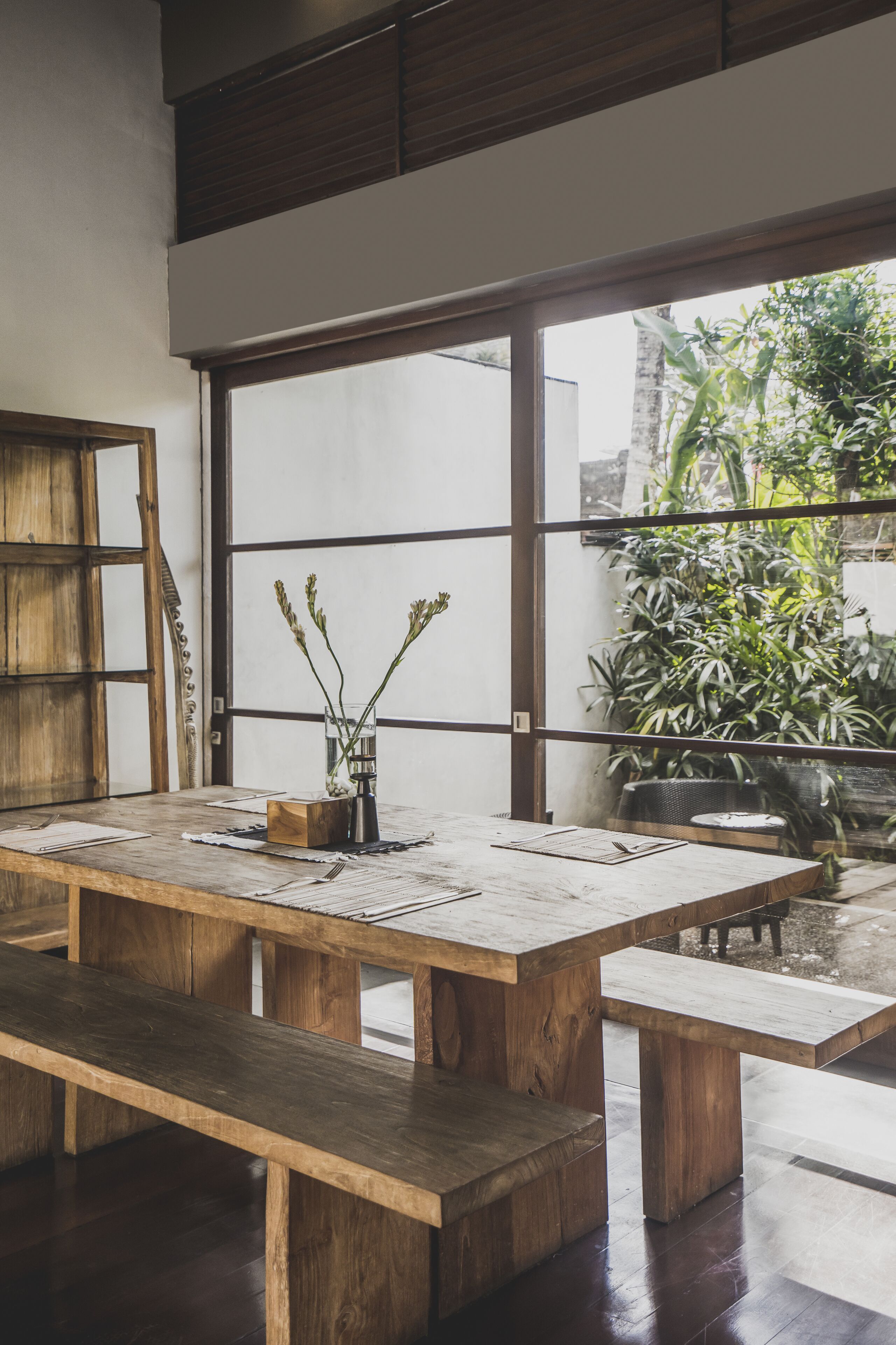 A wooden dining table with benches, a vase with greenery, and a view of a garden through large windows.