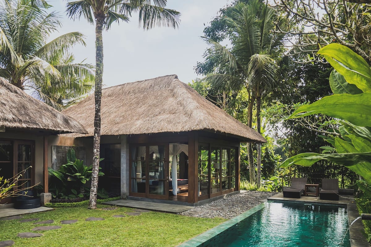 A pool area with a clear blue pool, a thatched-roof structure, and lush greenery.