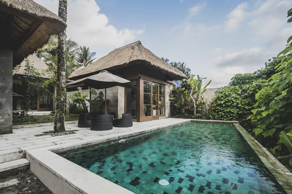 A pool area with a thatched-roof structure, surrounded by lush greenery and a clear pool.