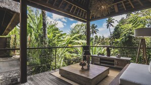 A wooden deck with a thatched roof, a wooden bench, a table with a bowl of fruit, and a lamp. - Luwak Ubud Villa & Jungle Suite (Ubud)