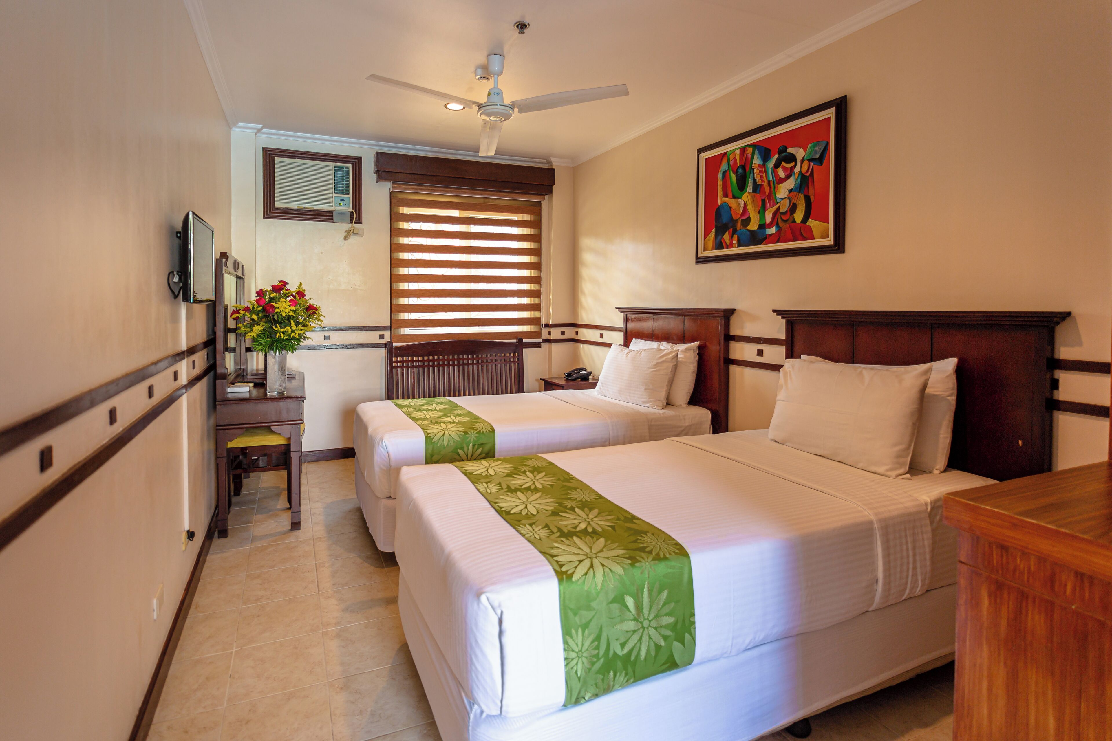 A hotel room with two beds, a desk, a chair, a television, a window with blinds, a ceiling fan, and a painting on the wall.