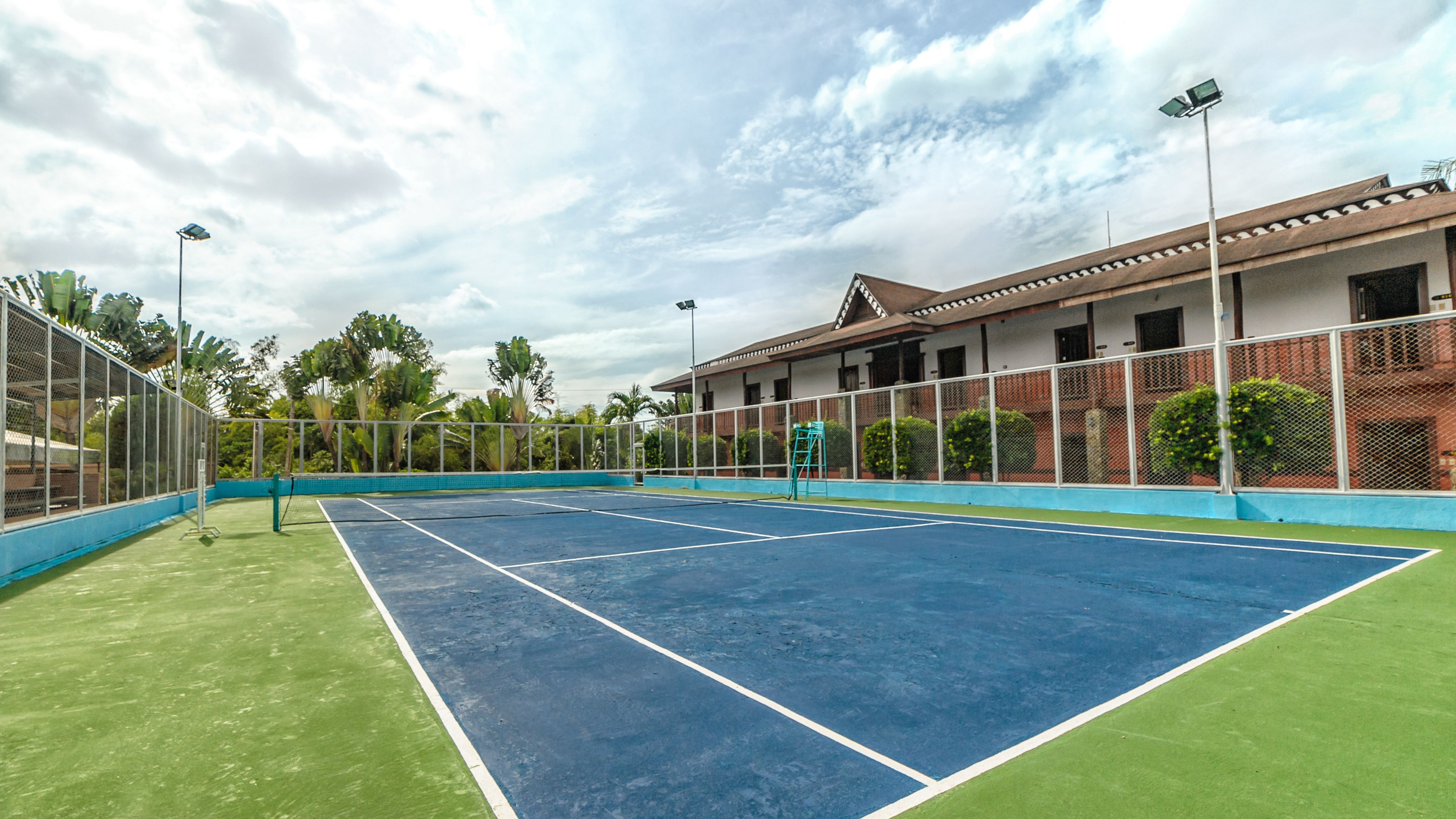 An outdoor tennis court with a net, surrounded by a fence and a building with a balcony.