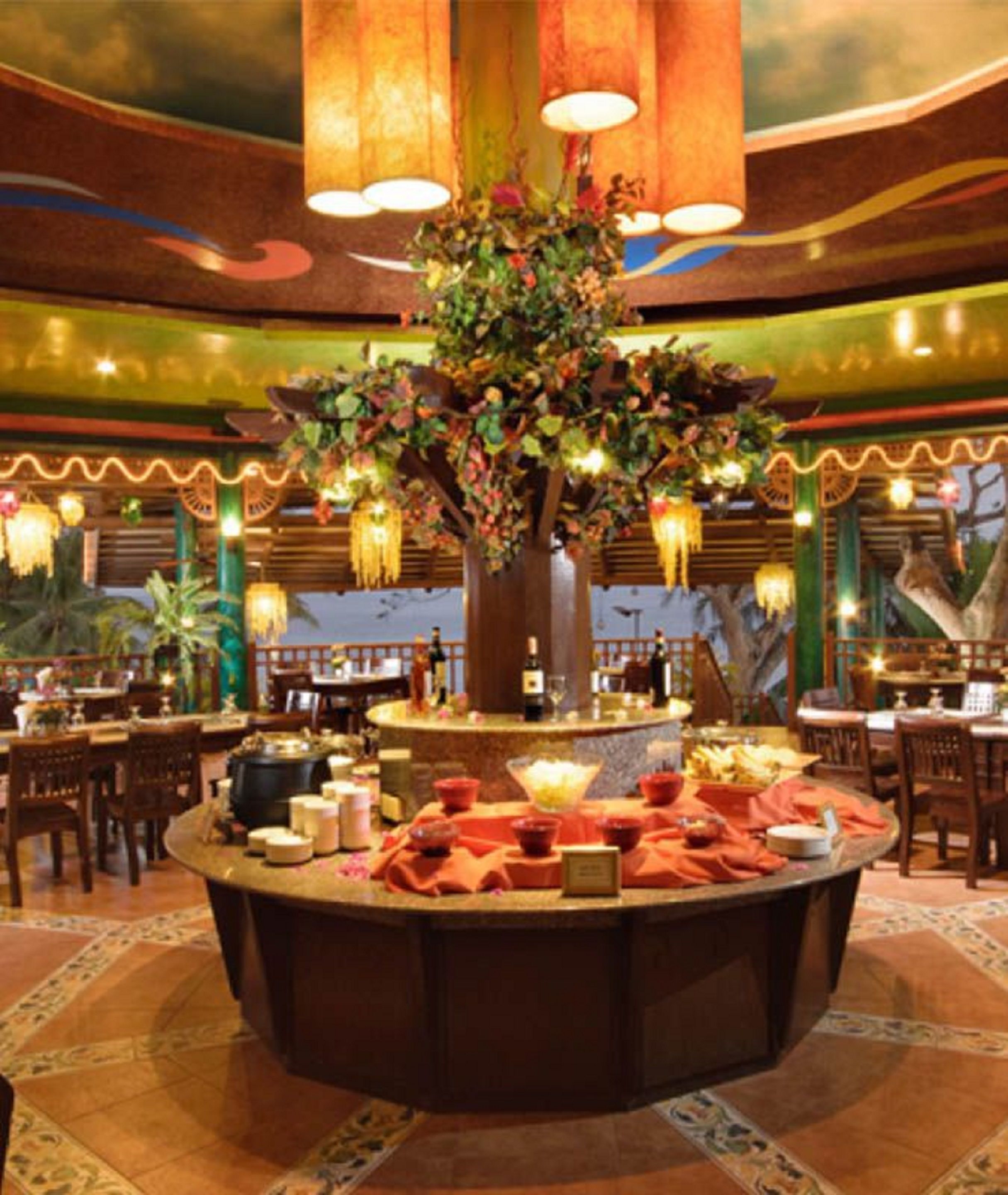 A restaurant with a central round table surrounded by chairs, a decorative tree centerpiece, and hanging lights.