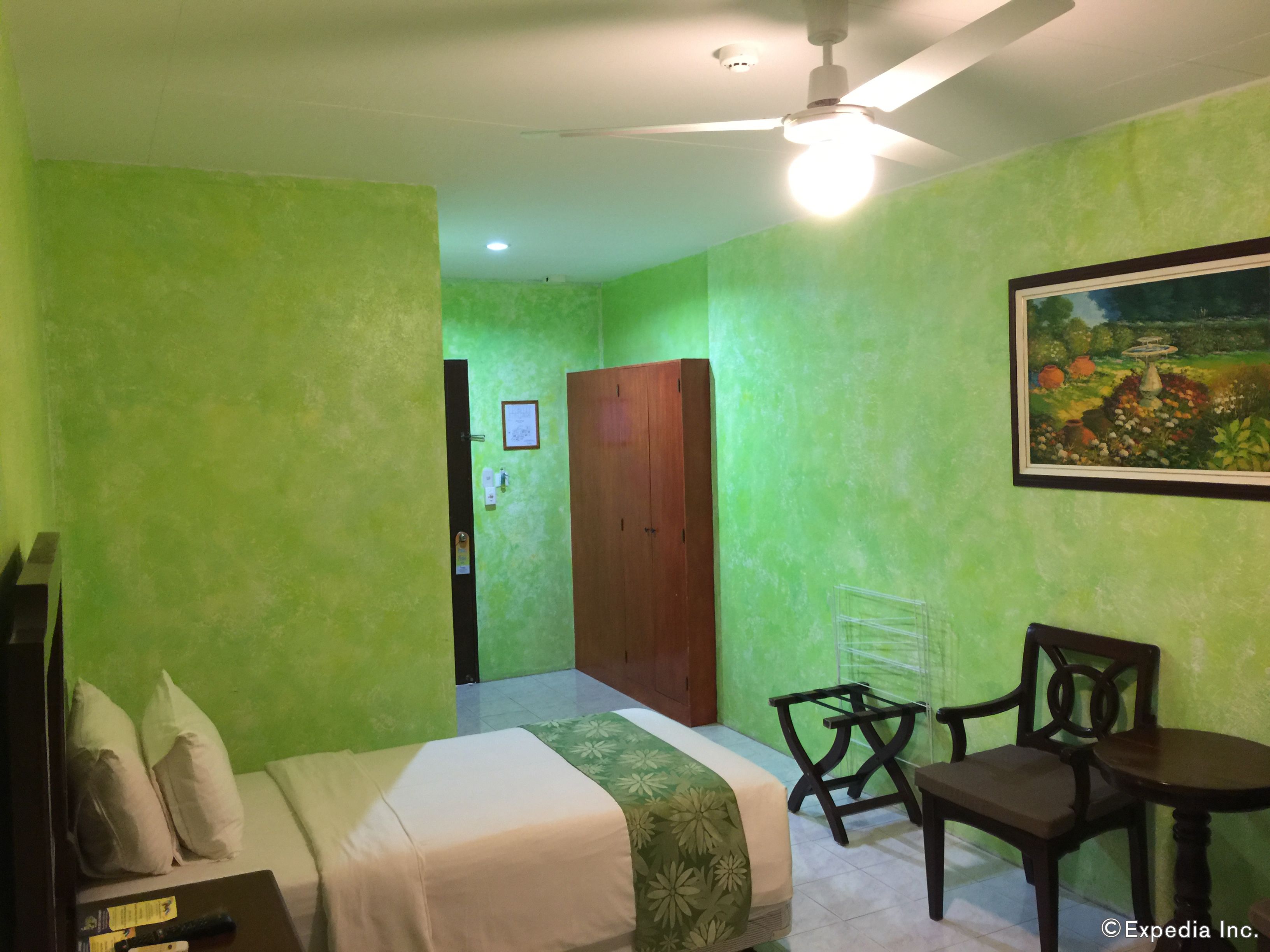 A room with green walls, a bed with a floral bedspread, a wooden door, a framed painting, a small table with a chair, and a ceiling fan.