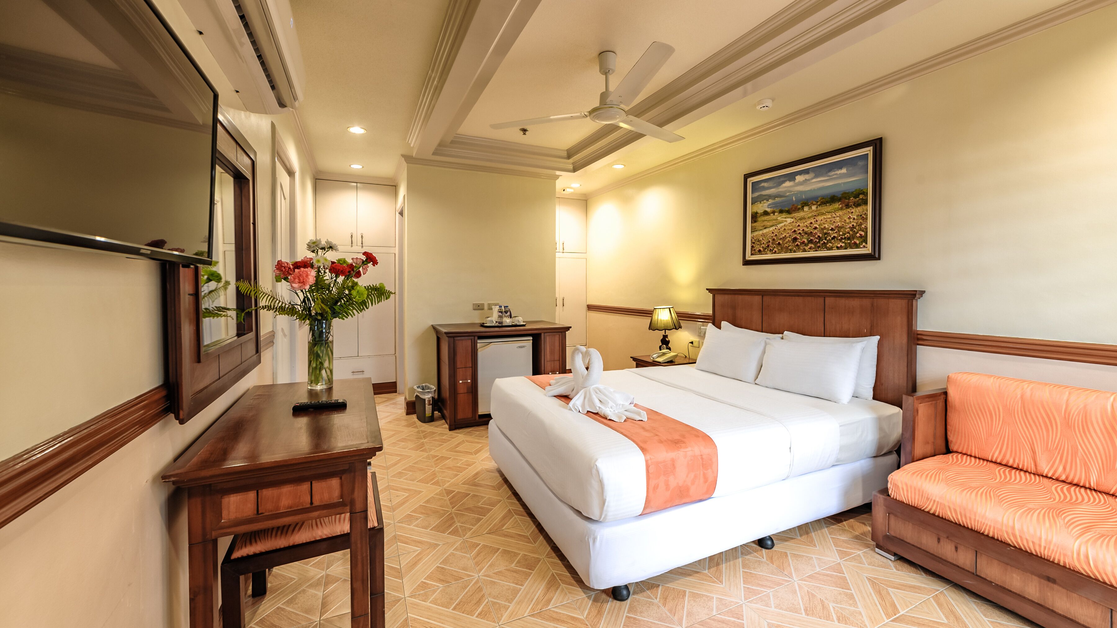 A hotel room with a large bed, a desk with a vase of flowers, a chair, a desk lamp, and a ceiling fan.