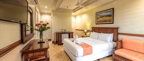 A hotel room with a large bed, a desk with a vase of flowers, a chair, a desk lamp, and a ceiling fan.