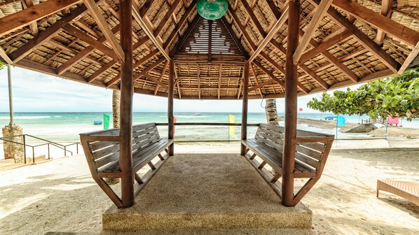 Un pabellón en la playa con techo de paja, dos bancos de madera y vista al océano.