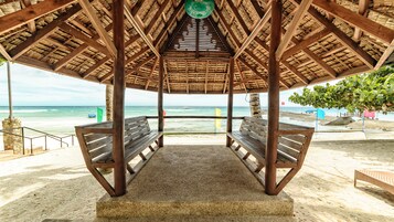 Un pabellón en la playa con techo de paja, dos bancos de madera y vista al océano.