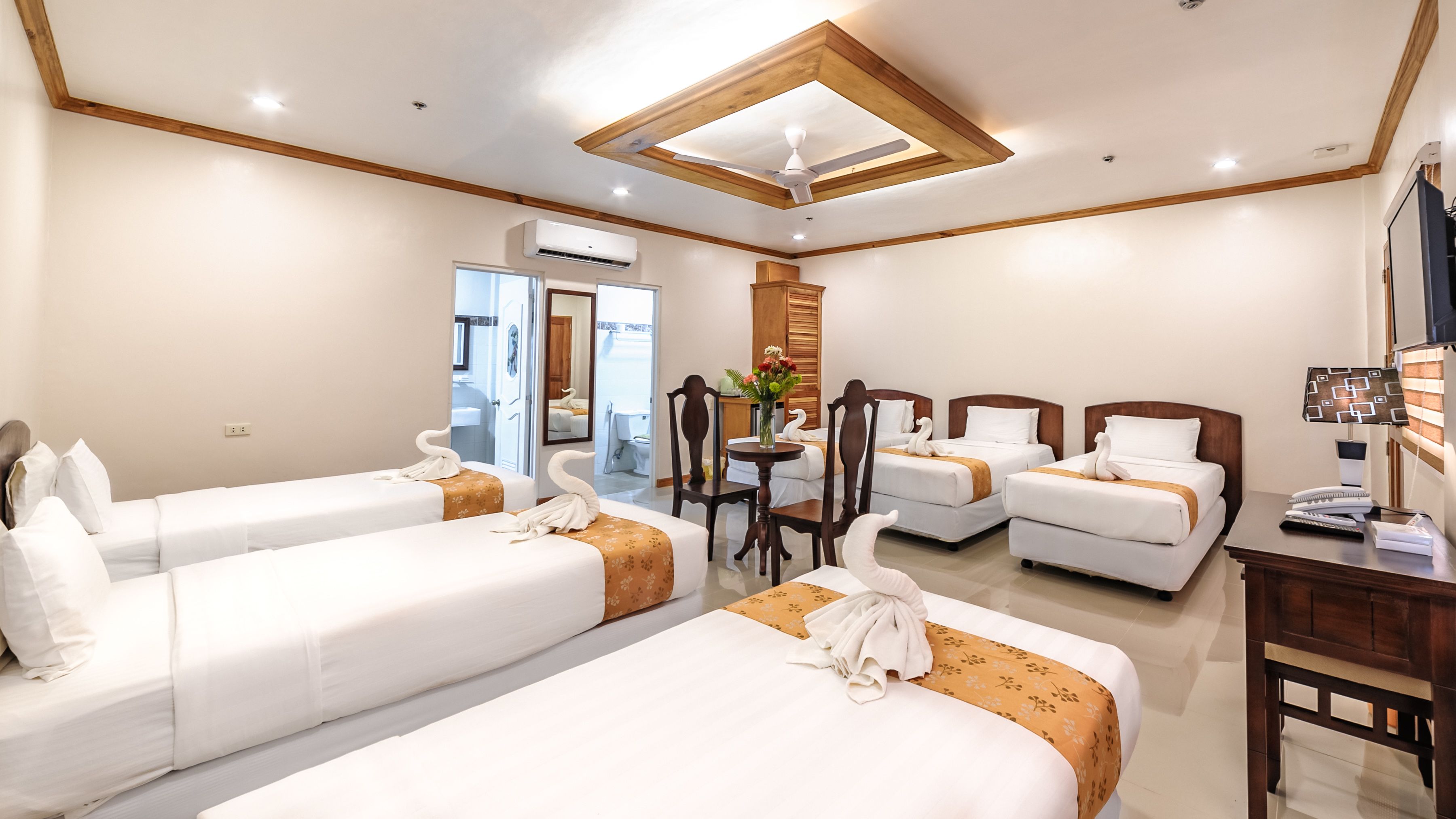 A hotel room with three single beds, a wooden ceiling, a desk with a lamp, and a mirror.
