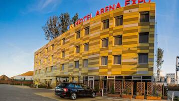 Un hotel llamado "Hotel Arena Expo" con fachada a cuadros, múltiples ventanas y un área de estacionamiento con un automóvil.